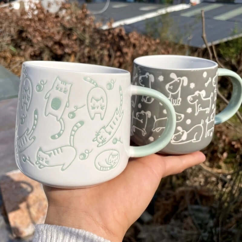 Two ceramic mugs featuring chubby cats and dogs, showcasing playful designs in a natural setting.