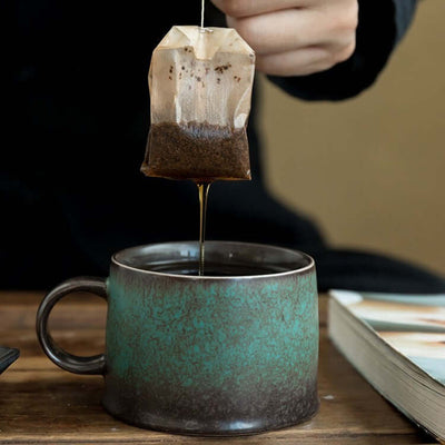 Person steeping tea in a peacock green retro ceramic mug with a teabag.
