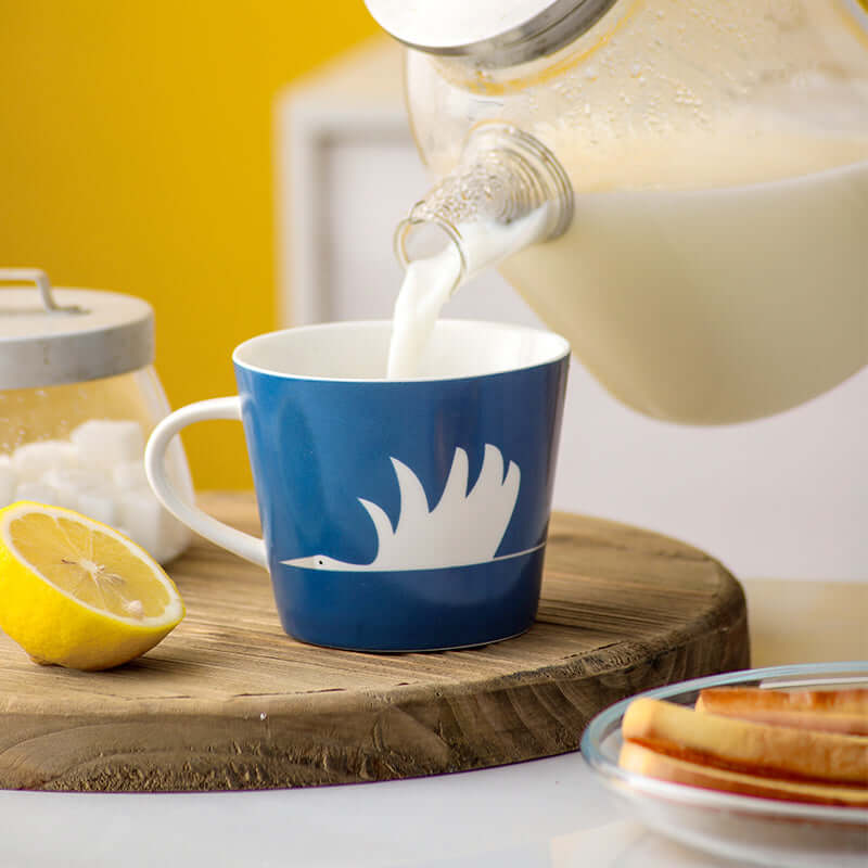 Ceramic blue mug featuring a white swan design, filled with milk, on a wooden board with lemon and snacks.
