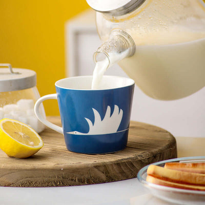 Ceramic blue mug featuring a white swan design, filled with milk, on a wooden board with lemon and snacks.