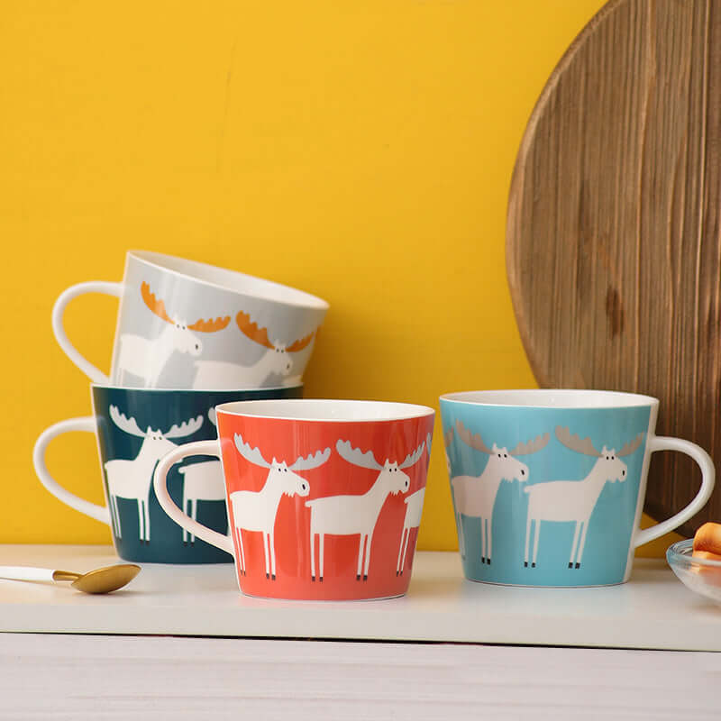 Colorful ceramic coffee mugs with moose design in red, teal, and blue against a bright yellow backdrop.