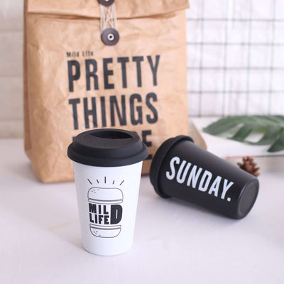 ROKEN stainless steel cups with lids, one white and one black, featuring playful designs, ideal for coffee or tea.